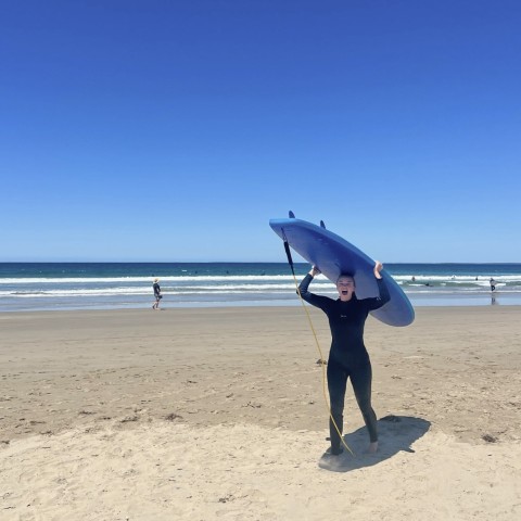 Student on the beach wears a wetsuit and carries a surfboard over her head.